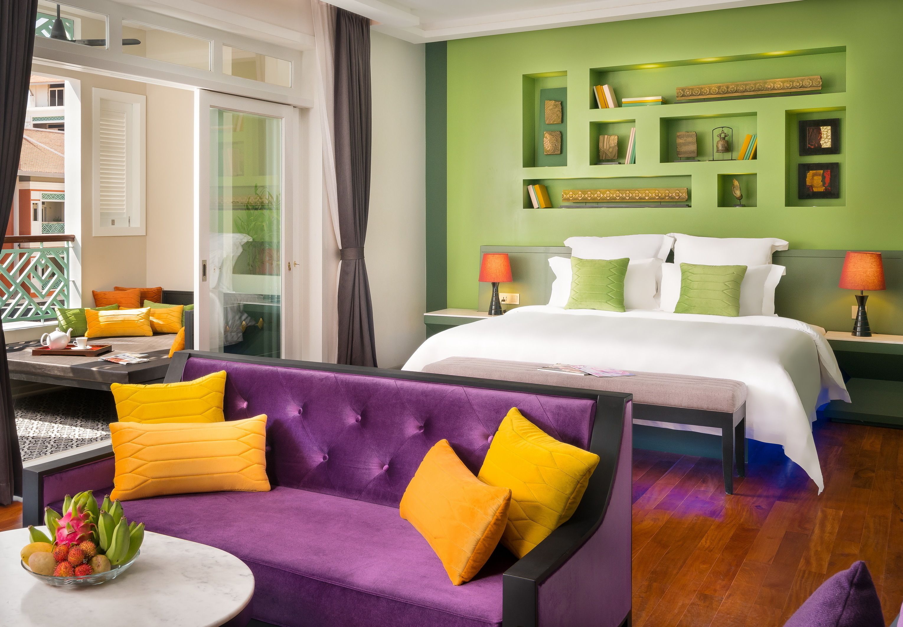 Ein Hotelzimmer mit einem violetten Sofa, einem Bett mit weißer und grüner Bettwäsche, einem kleinen Tisch mit Obst und einer grünen Akzentwand mit Regalen.