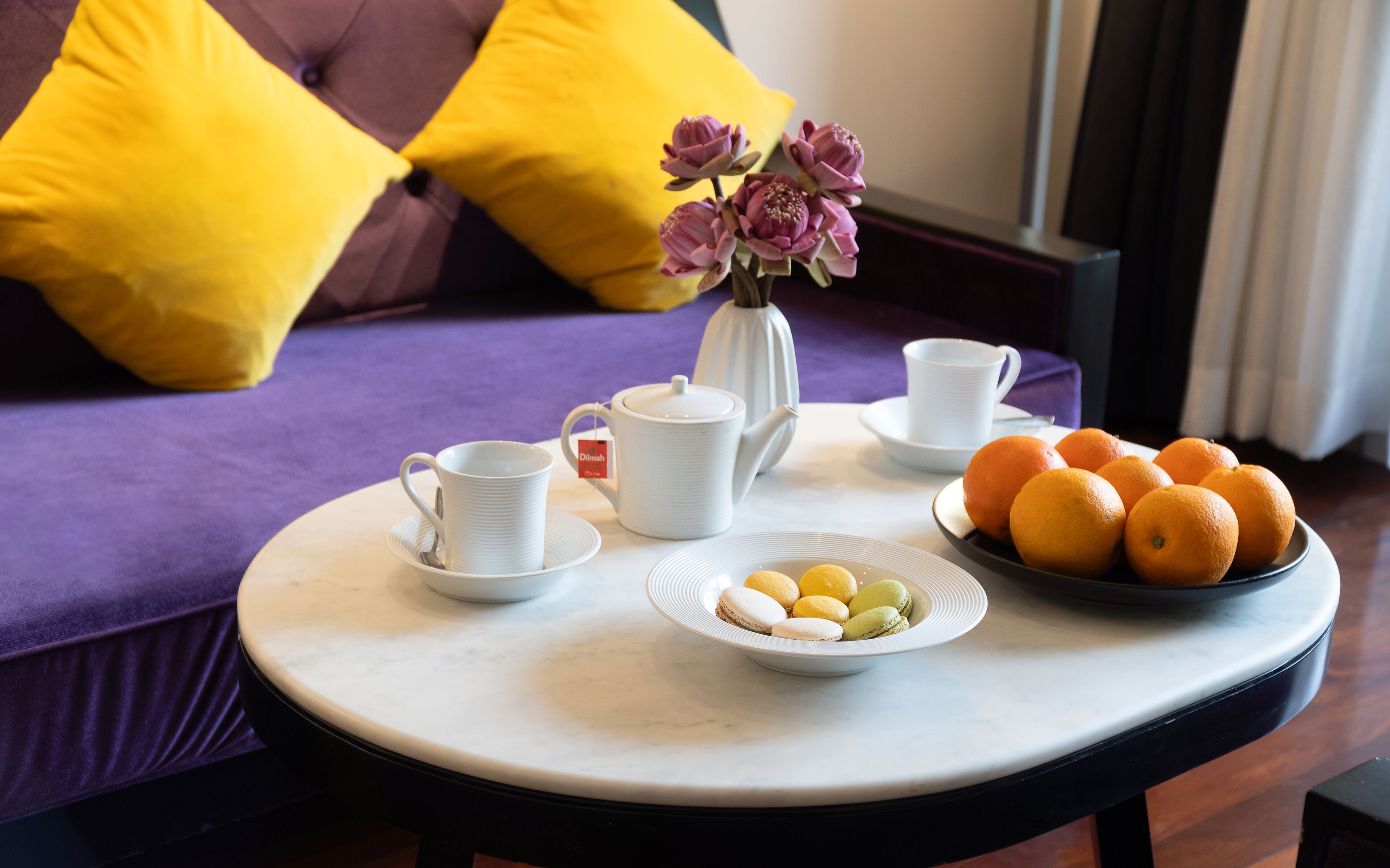 Una mesa con un calentador de agua, tazas, un plato de naranjas y un plato de macarons.