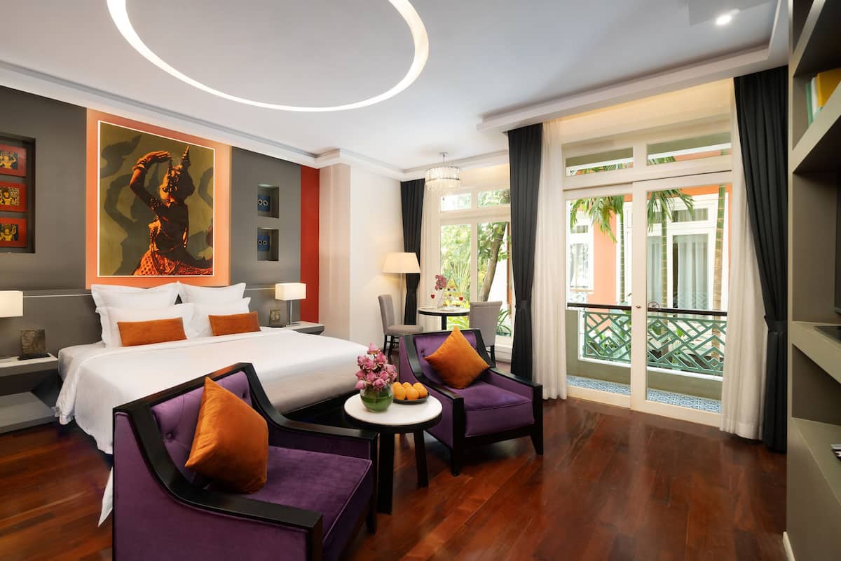 Ein Hotelzimmer mit einem Bett, zwei violetten Sesseln, einem kleinen Tisch mit Blumen und einem großen Fenster mit Blick auf Grünflächen.