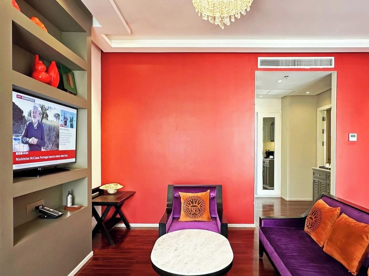 A modern living room with a red accent wall, a television, a round white ottoman, a purple sofa, and a small table with a lamp.