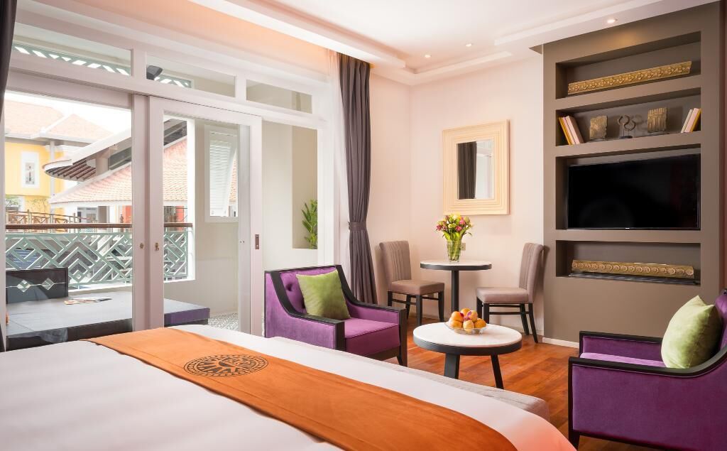 A hotel room with a bed, two purple armchairs, a small round table with fruit, a TV, and a balcony with a view.