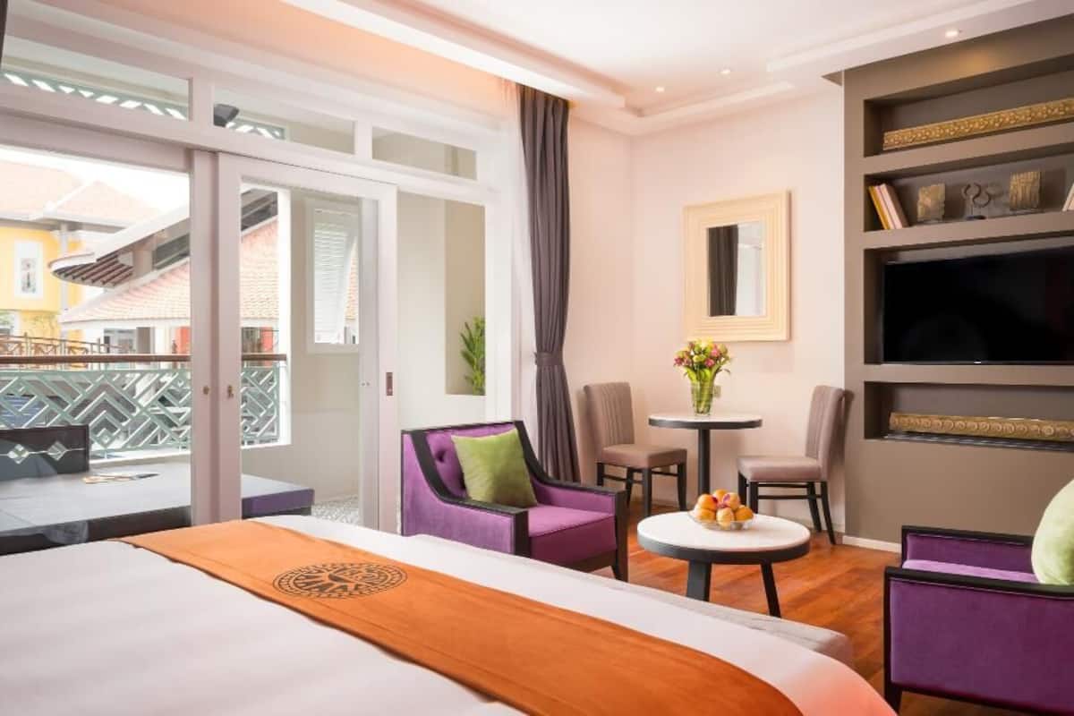 Ein Hotelzimmer mit einem Bett, zwei violetten Sesseln, einem kleinen runden Tisch mit Obst, einem Fernseher und einem Balkon mit Blick.