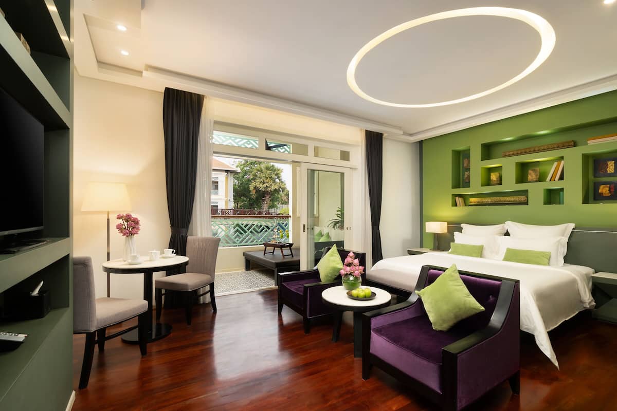 Ein Hotelzimmer mit einem Bett, zwei violetten Sesseln, einem kleinen Tisch mit einer Vase, einem Fernseher und einem Balkon mit Blick auf Grünflächen.