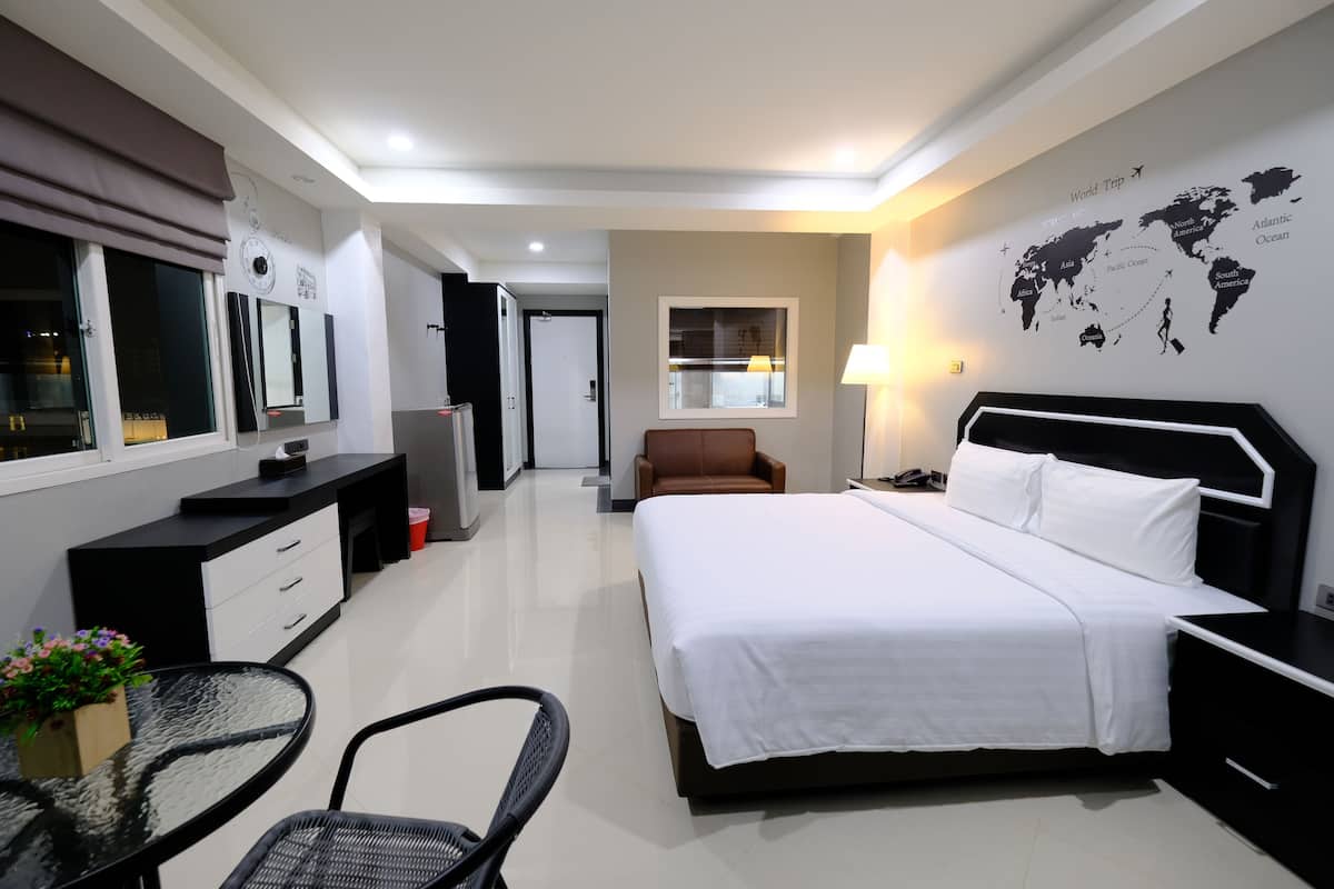 A modern hotel room with a large bed, a desk with drawers, a chair, a small table with flowers, a lamp, and a world map on the wall.