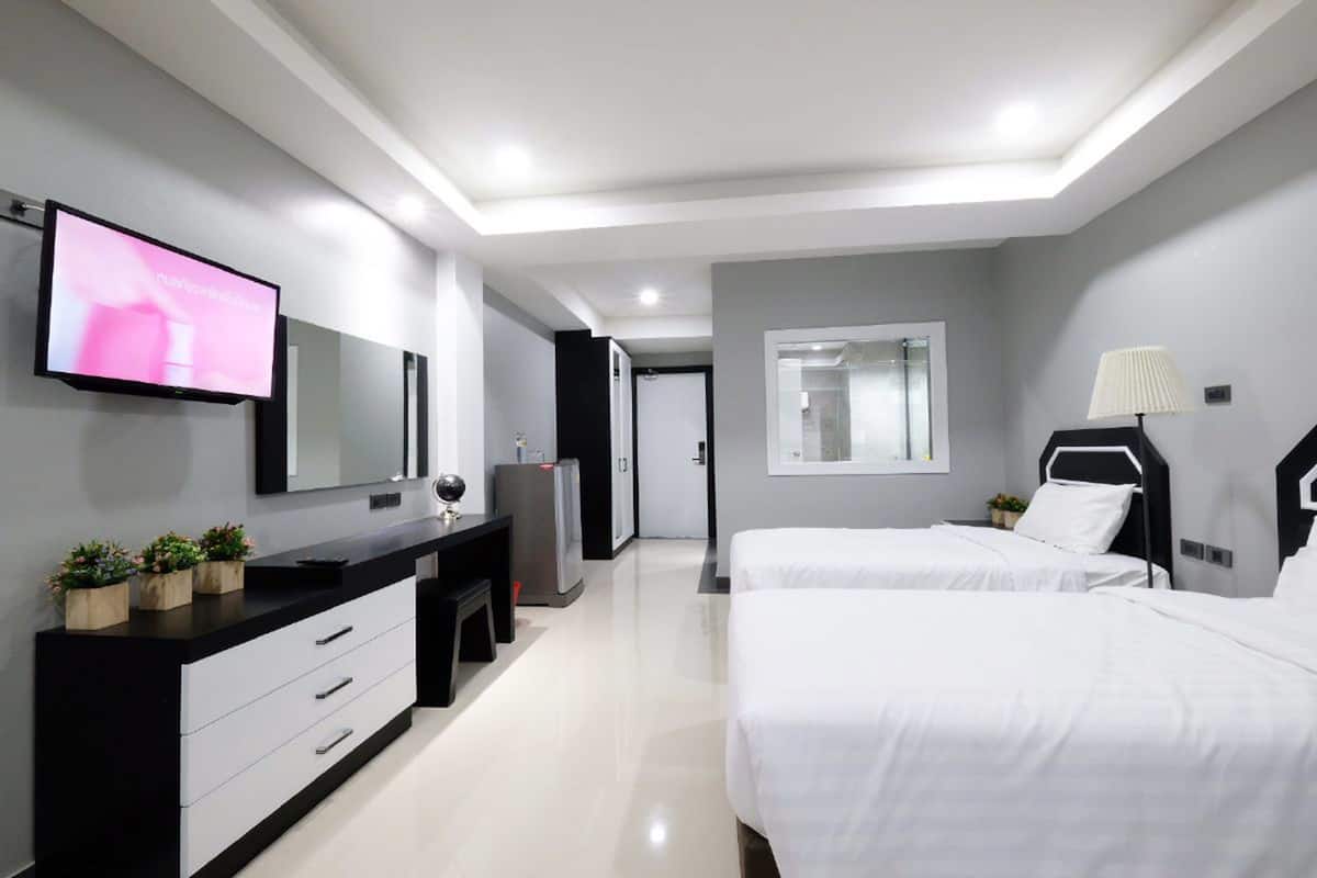A modern hotel room with a large flat-screen TV mounted on the wall, a black and white vanity, two beds with white linens, and a large mirror.