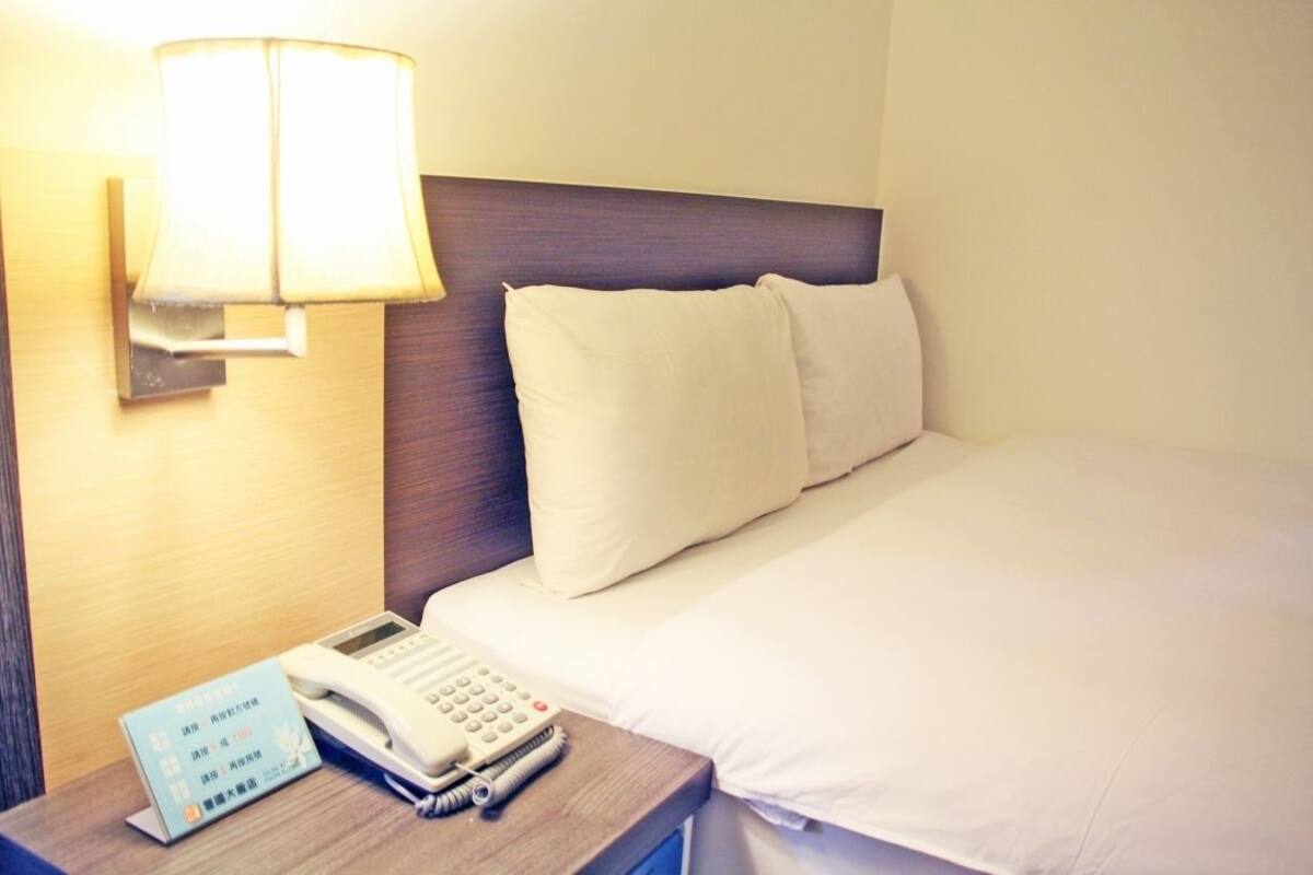 A hotel room with a bed, a bedside table with a lamp, a phone, and a sign with room information.