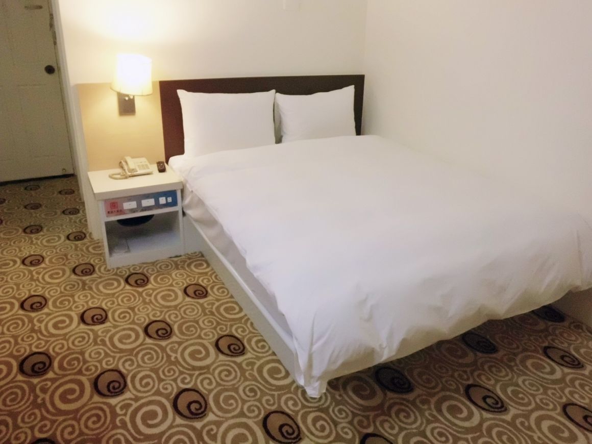 A hotel room with a bed, a nightstand with a lamp, and a patterned carpet.