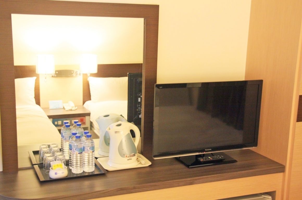 A hotel room with a bed, bedside lamps, a flat-screen TV, a tray with bottled water, a kettle, and a remote control.