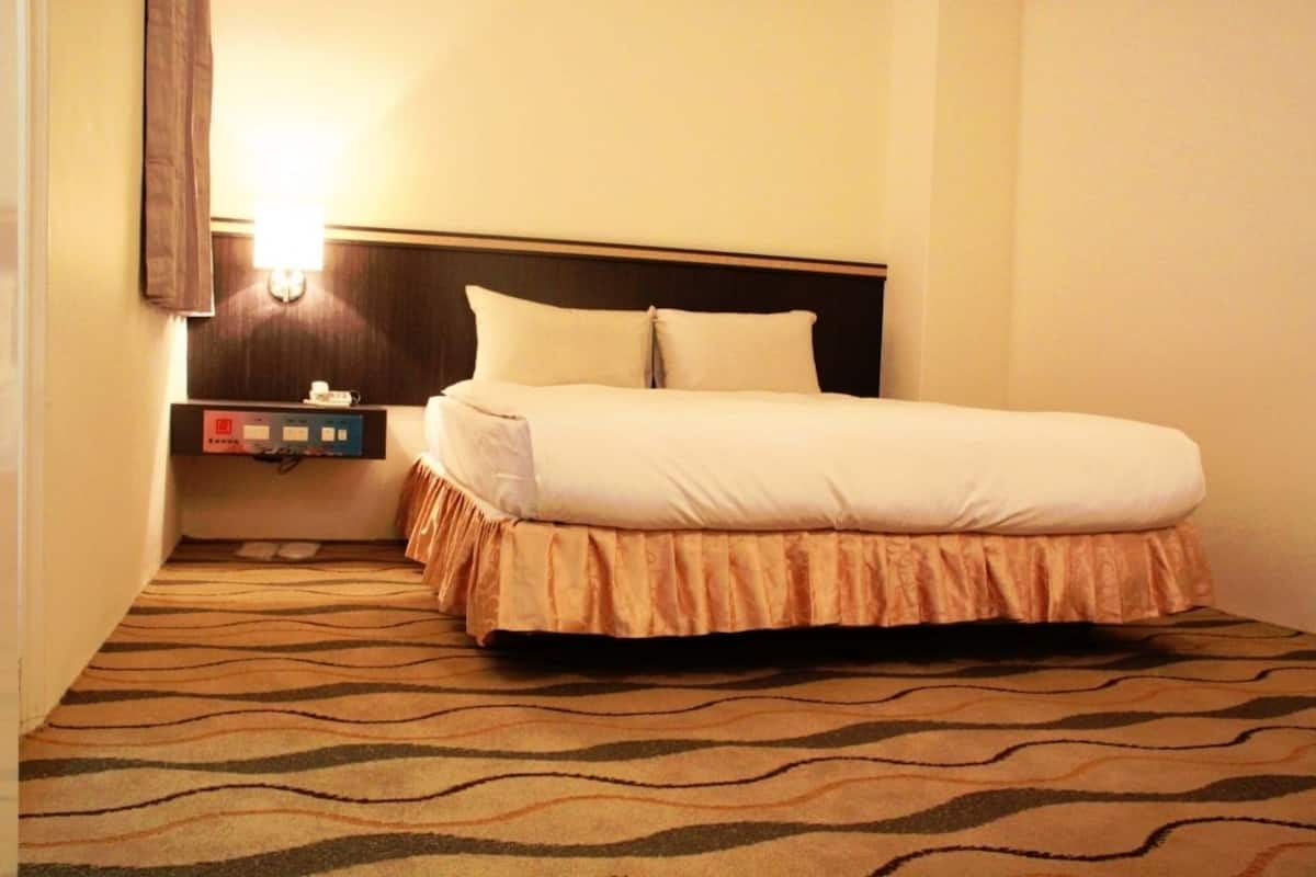 A hotel room with a bed, a control panel, and a patterned carpet.