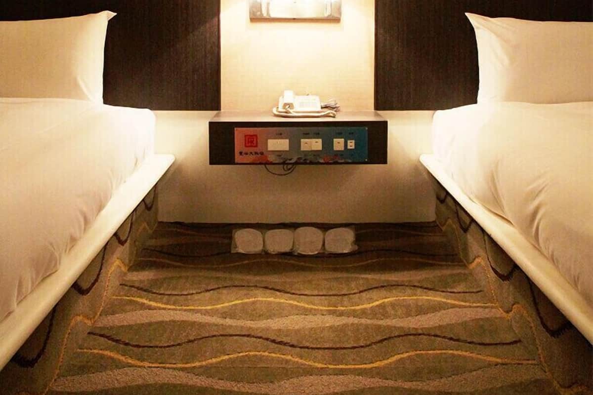 A hotel room with two beds, a nightstand with a lamp, and a control panel.