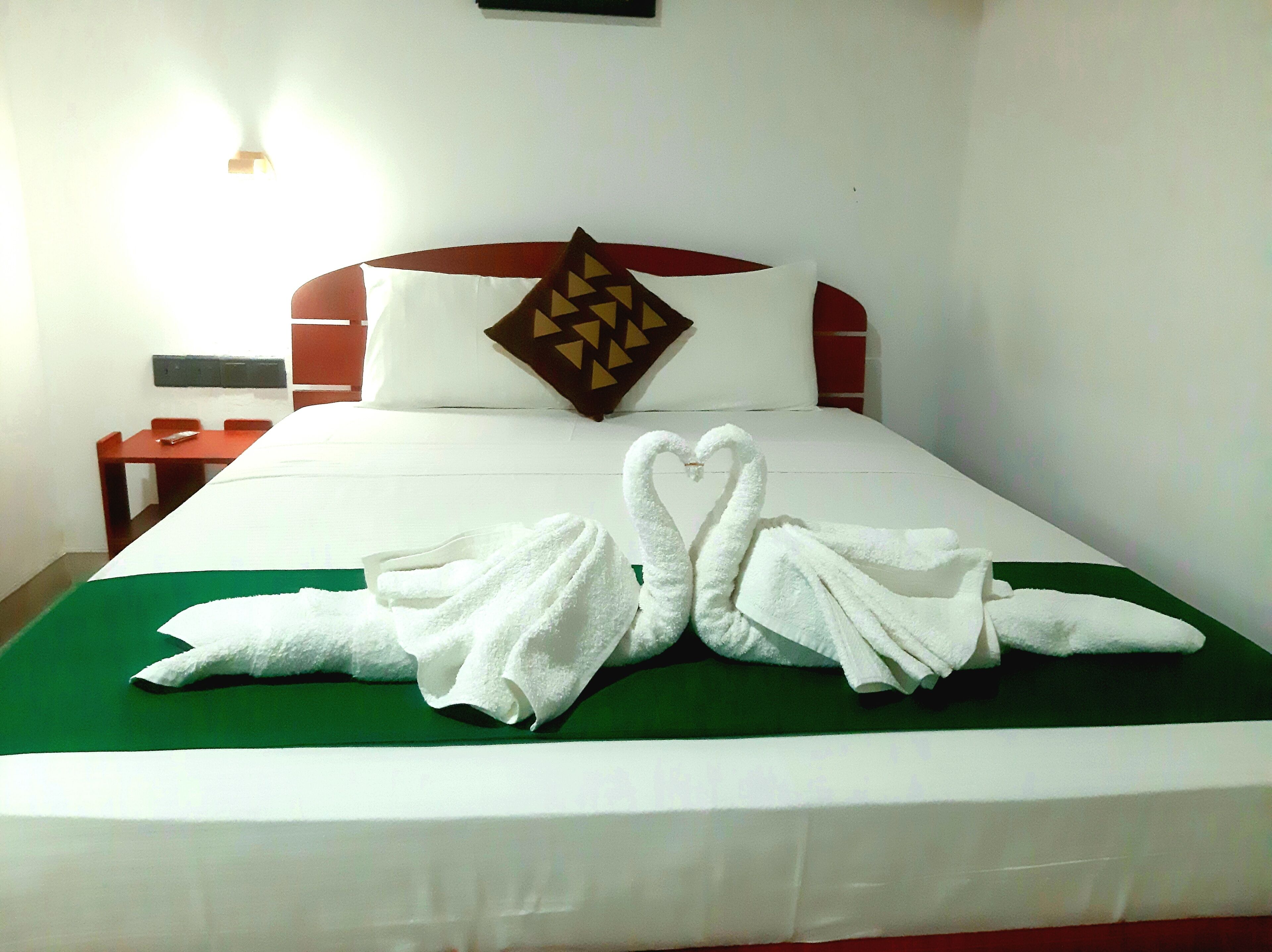 Quarto de hotel com cama arrumada, manta verde e duas toalhas dispostas em formato de coração.