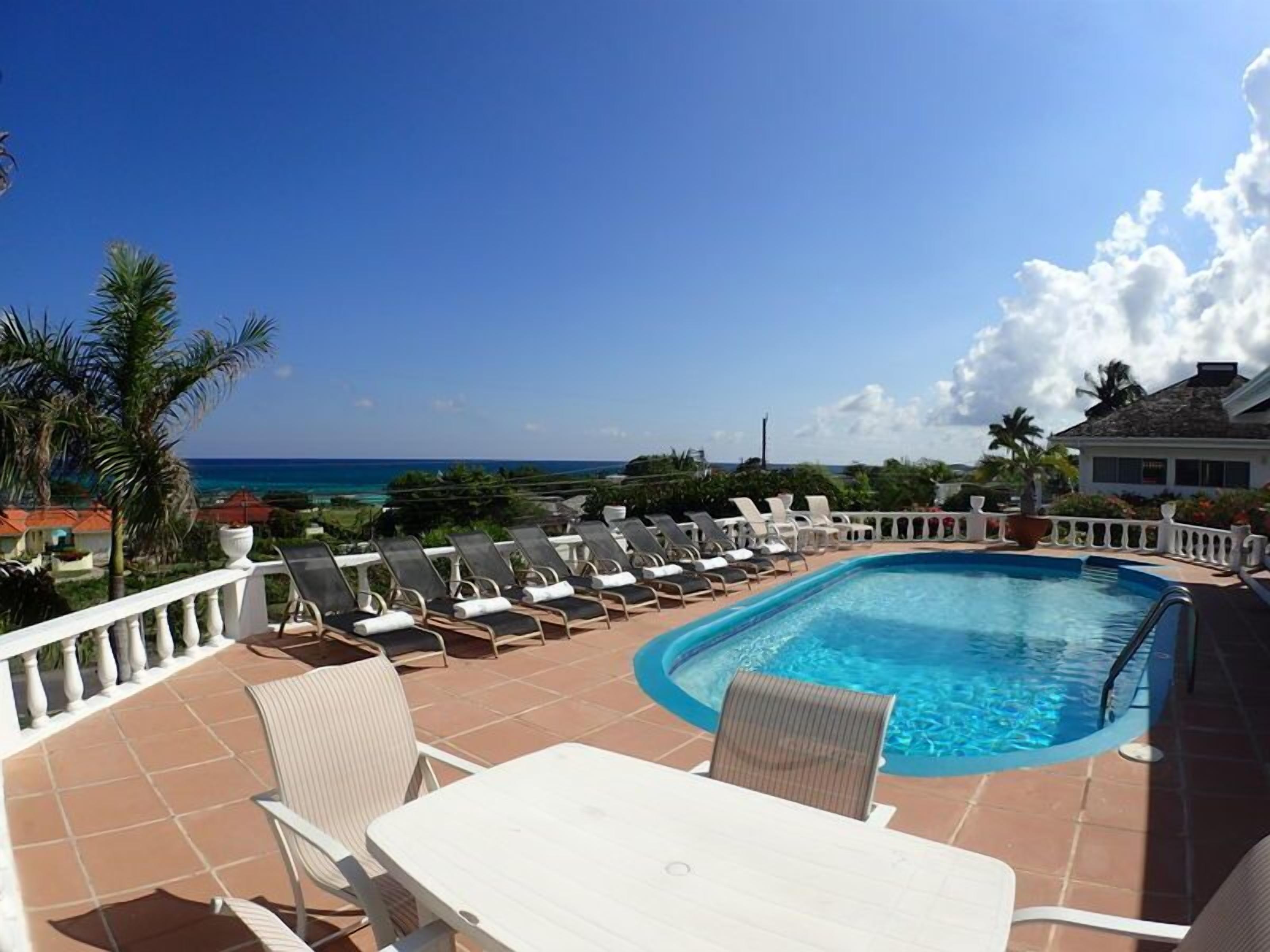 A pool area with lounge chairs and a table, overlooking a beach and ocean.