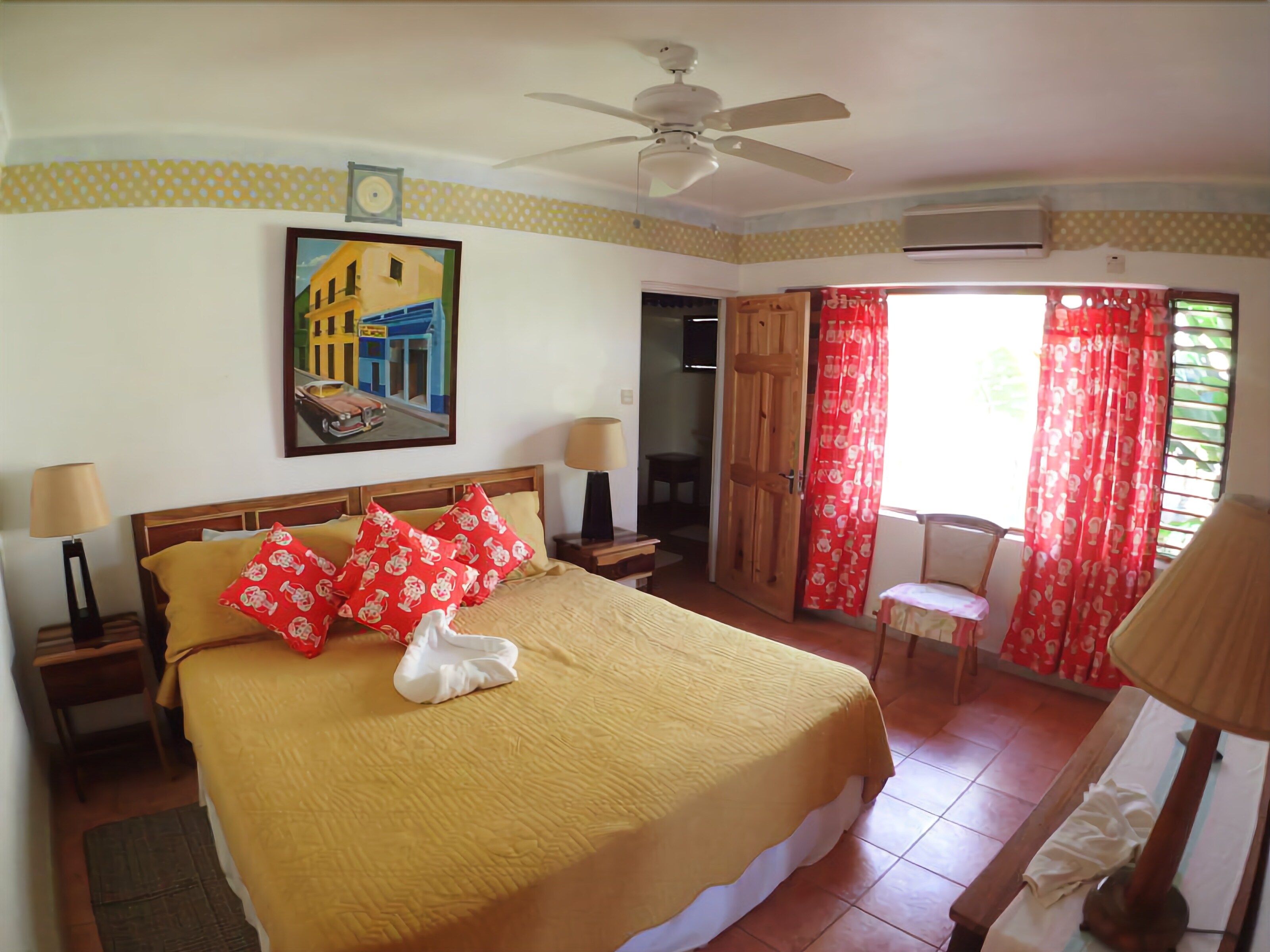 A hotel room with a bed, two bedside tables, a ceiling fan, a painting on the wall, and a window with red curtains.