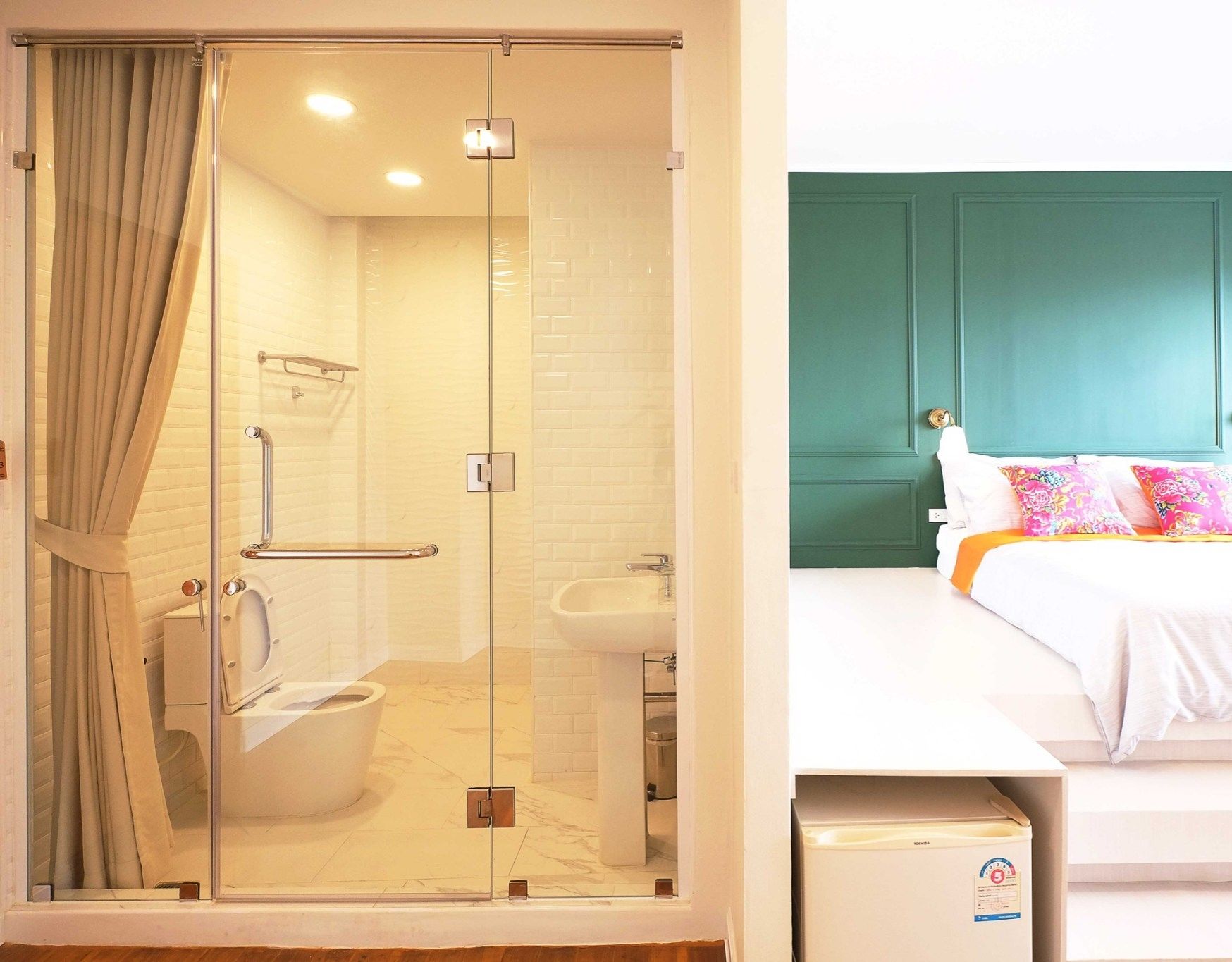 A hotel room with a glass-enclosed shower, a toilet, a sink, and a bed with a teal headboard and colorful pillows.