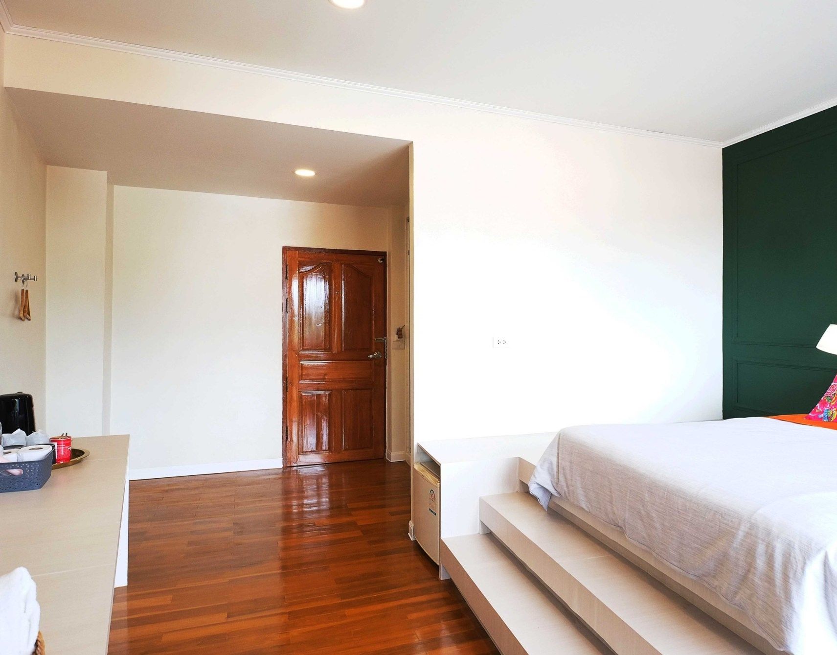 Habitación de hotel con piso de madera, cama con colchón blanco, pared verde decorativa y un escritorio con lámpara.