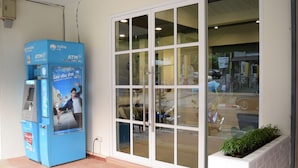 An ATM machine with a poster advertising a mobile service, a large glass door, and a planter with greenery. - Buakham Rim Khong (Chiang Saen)