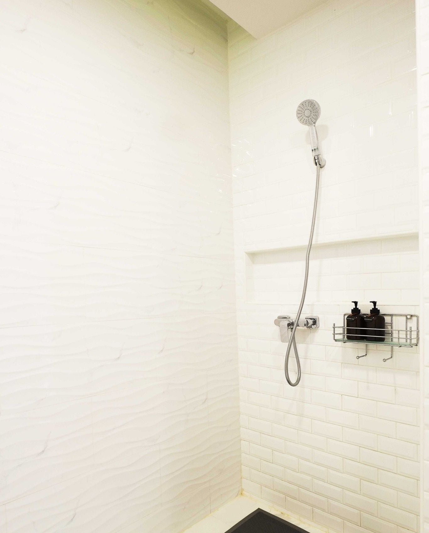 A shower with a handheld showerhead and a wall-mounted faucet. There is a metal shelf with two bottles of shampoo and conditioner.