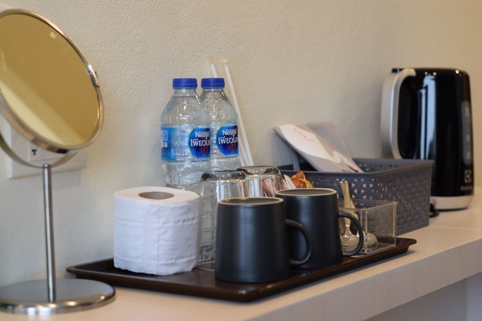 A hotel reception area with a mirror, two Nestle water bottles, a coffee pot, a tray with a roll of toilet paper, and a black mug.