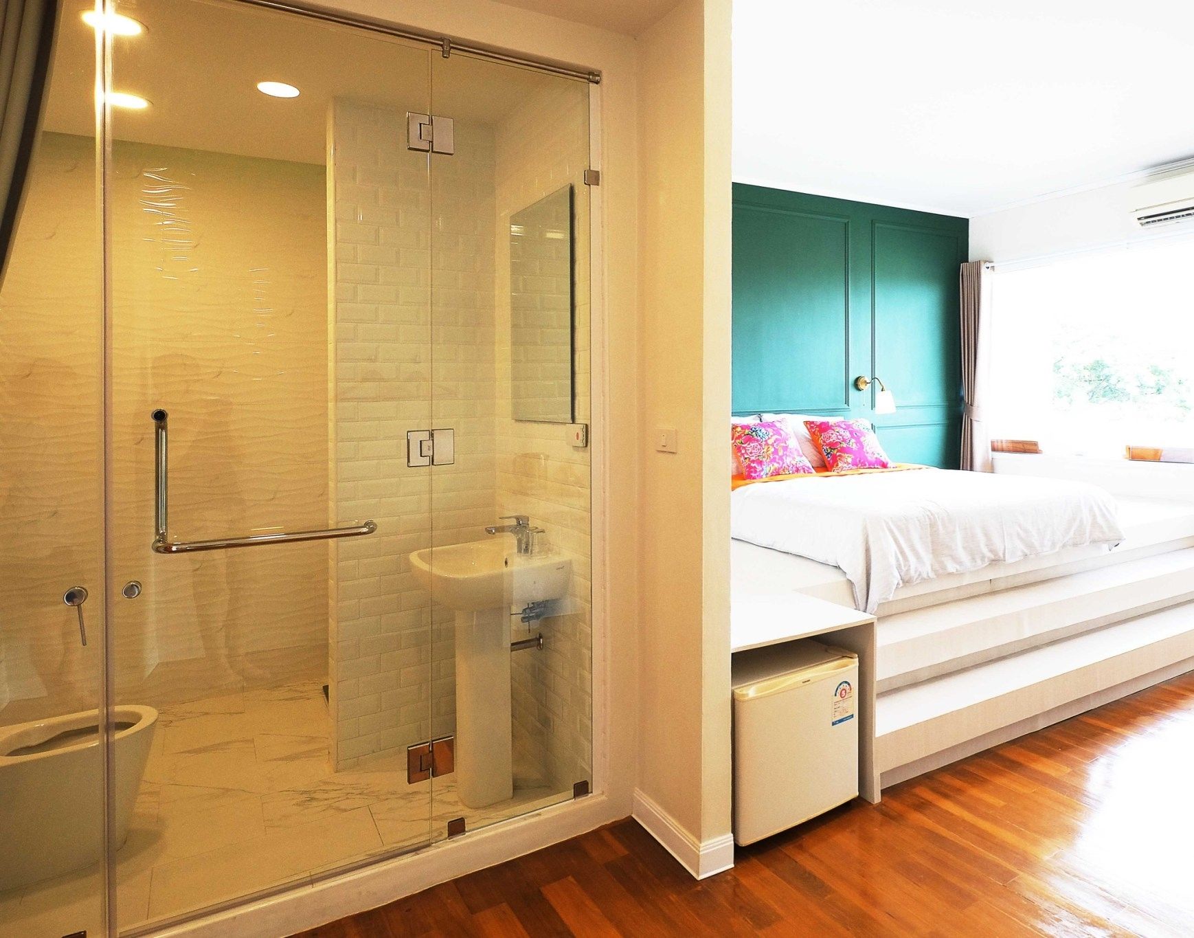 A hotel room with a glass-enclosed shower, a white sink, a bed with a teal headboard, and a window with curtains.