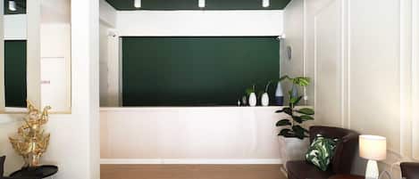 A reception area with a green backdrop, a reception desk, a brown leather sofa, a small table with a lamp, and a decorative golden statue.