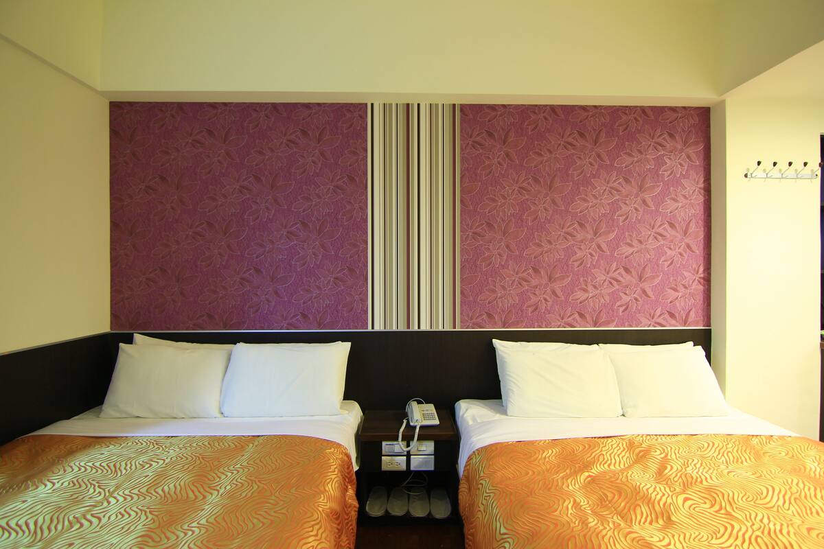 A hotel room with two beds, a nightstand with a phone, and a shoe rack under the beds.