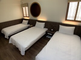 Habitación de hotel con tres camas individuales, piso de madera, espejo redondo y una mesita de noche pequeña con un teléfono y una caja de pañuelos.