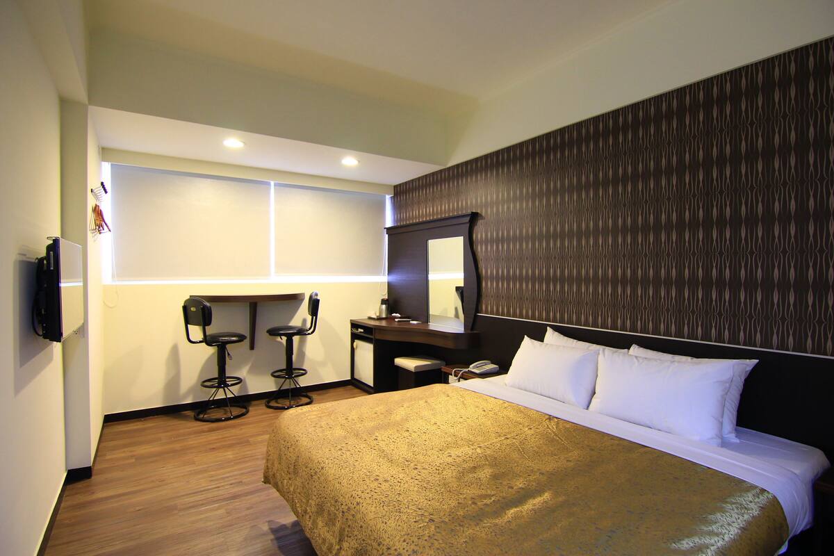 A hotel room with a large bed, a desk with a chair, a mirror, and a TV mounted on the wall.