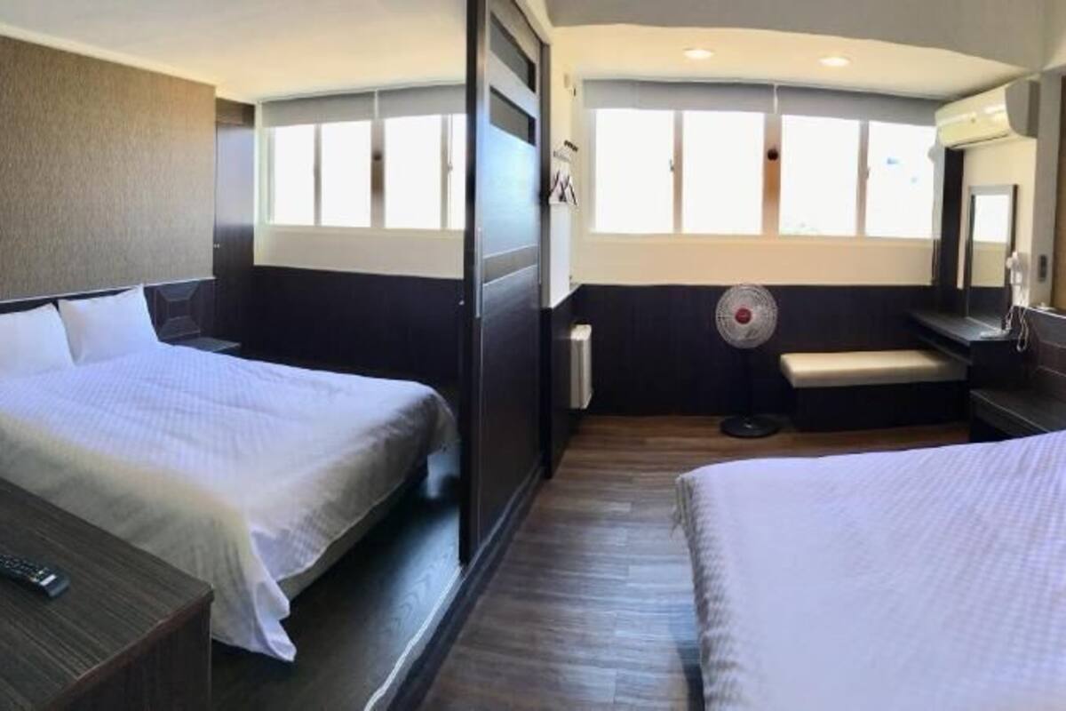 A hotel room with two beds, a bench, a fan, and a large window.