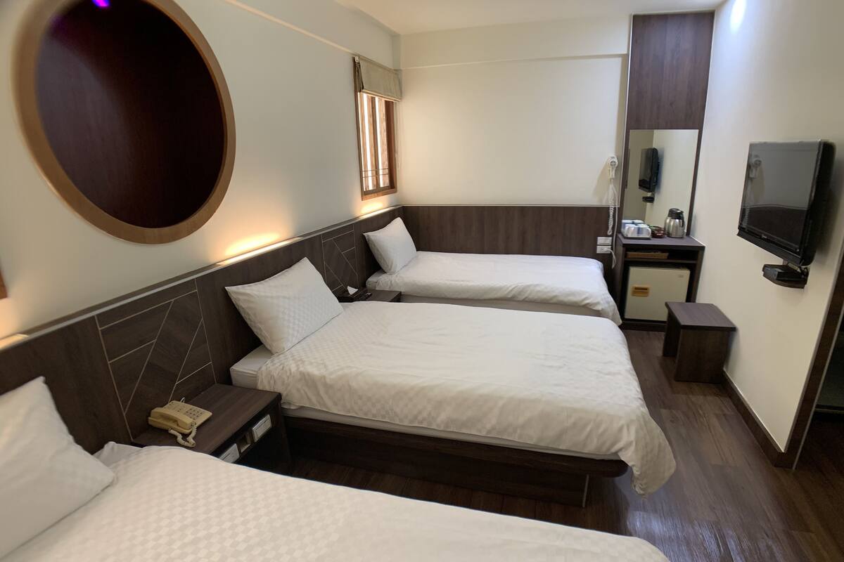 A hotel room with two beds, a desk, a TV, and a small kitchenette.