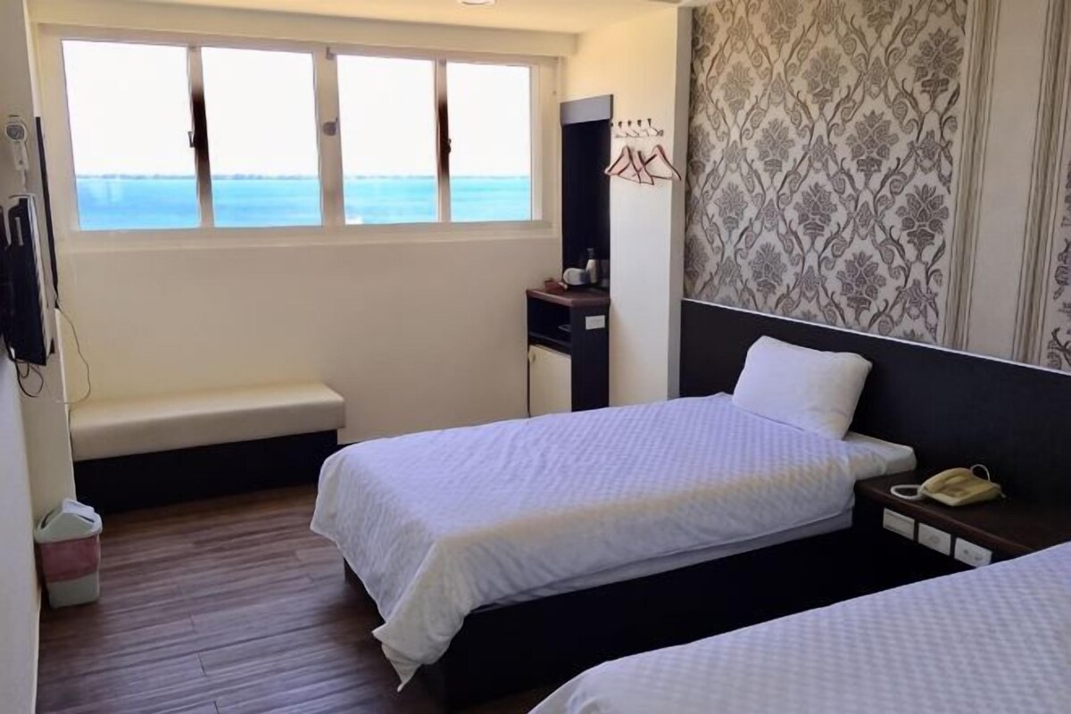 A hotel room with two beds, a bench, a desk, a telephone, and a window with a view.