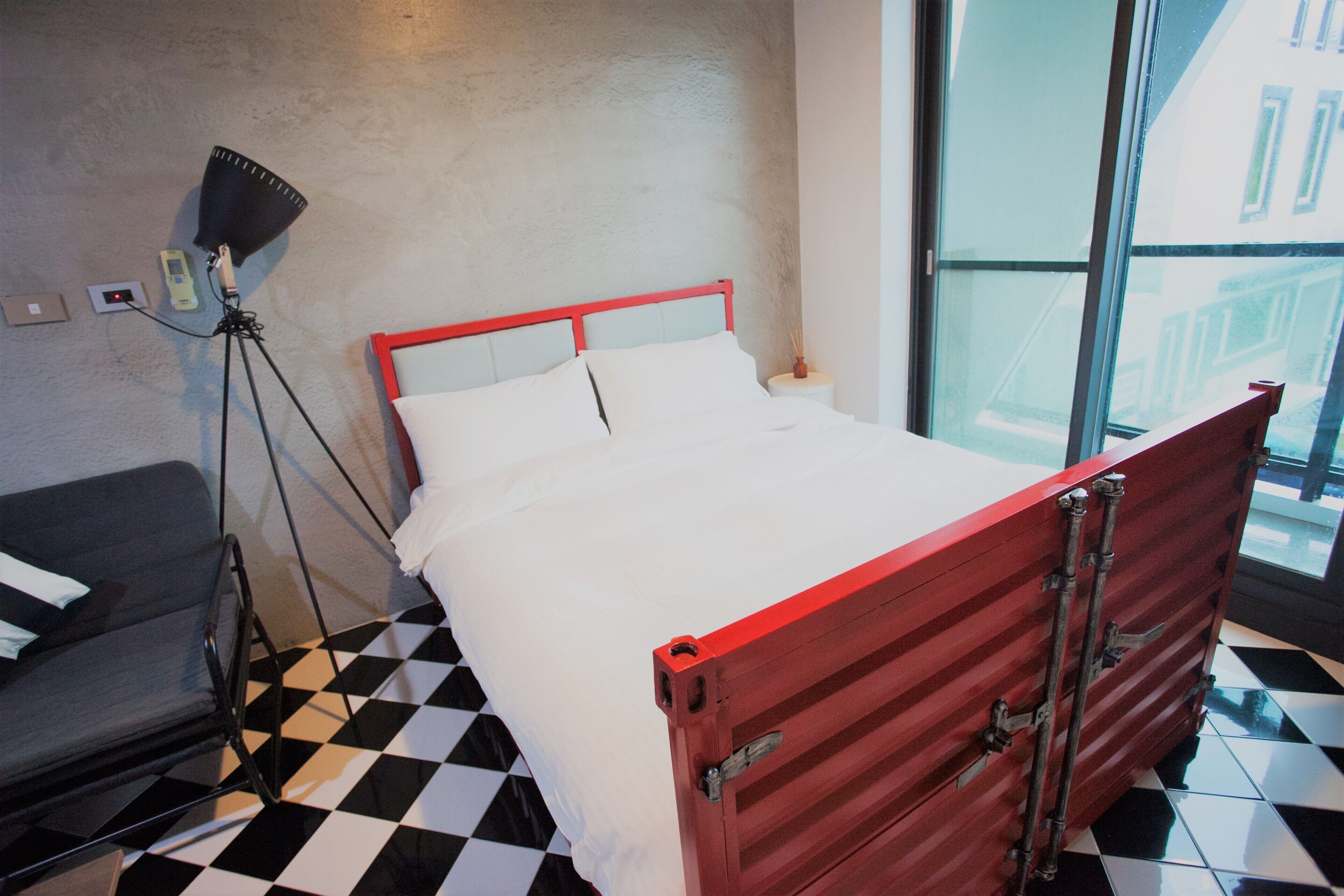 A bed with a red frame, a gray chair, and a floor lamp in a room with a checkered floor.