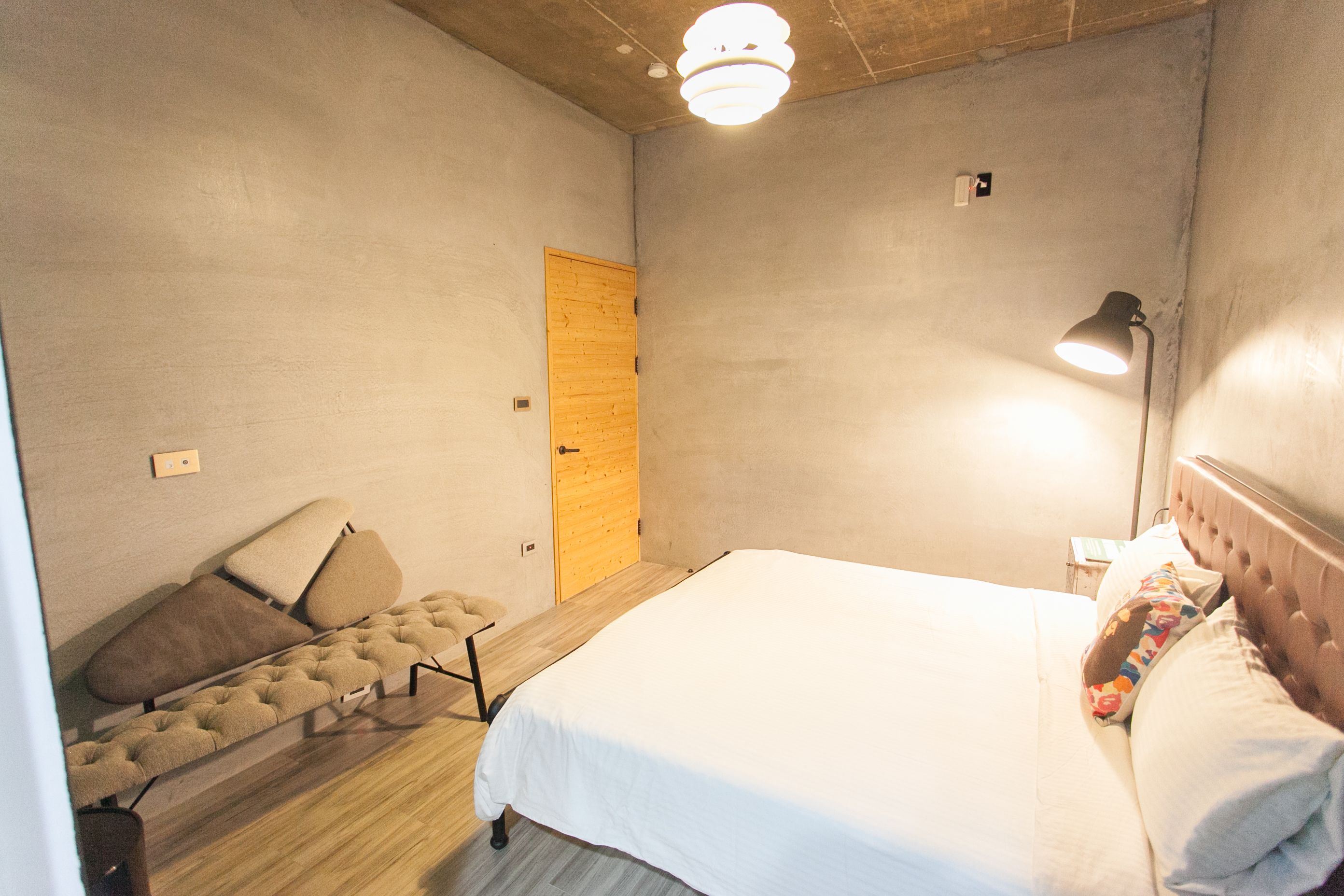 Un dormitorio con una cama, una puerta de madera, una lámpara de noche y una pequeña zona de estar con cojines.