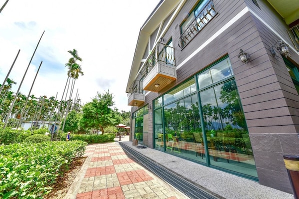 A modern building with large glass windows, a paved walkway, and a garden area with palm trees.