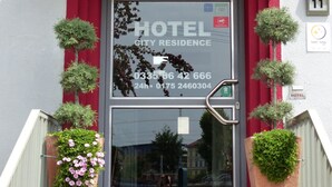 Entrada de hotel con toldo rojo, puerta de cristal y dos macetas a cada lado.