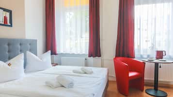 A hotel room with a bed, two pillows, a red chair, a small table with a kettle, and a window with curtains.