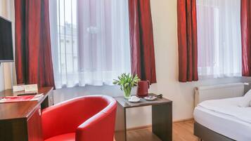 A hotel room with a bed, a desk with a chair, a small table with a plant and a red mug, and red curtains on the windows.