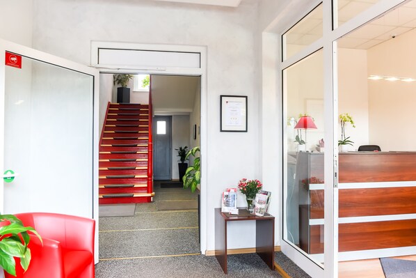 A reception area with a red chair, a wooden desk, a framed certificate, and a small table with a plant and flowers. - Hotel City Residence Frankfurt-Oder (Frankfurt an der Oder)