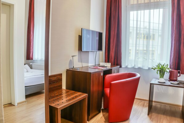Una habitación de hotel con cama, un escritorio con televisor, una silla roja, una mesita con planta y taza, y una ventana con cortinas.