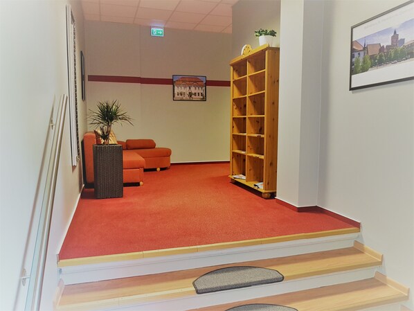 Zona de recepción con alfombra roja, estantería de madera, un cuadro enmarcado y una planta.