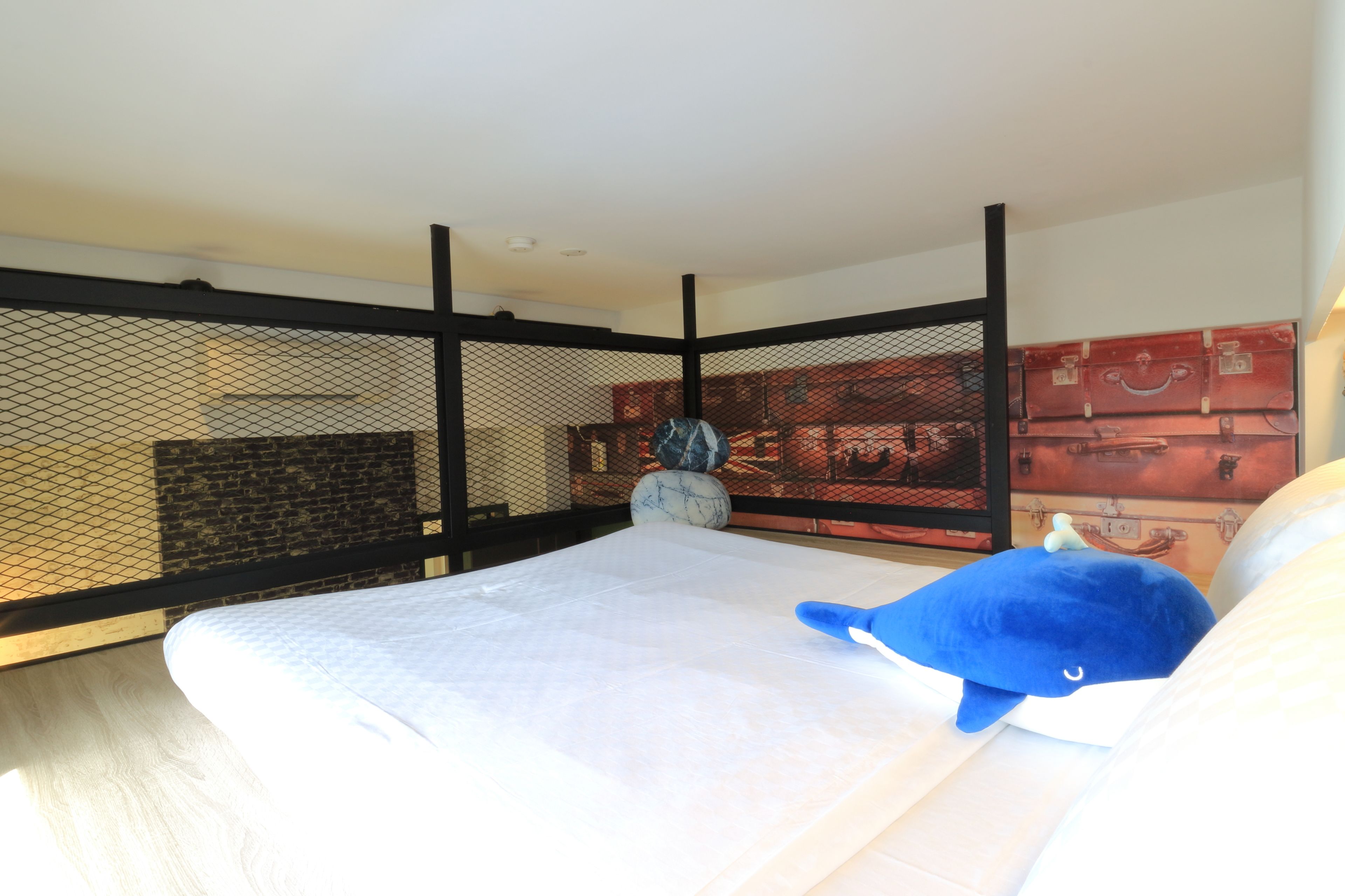 Une chambre à coucher avec un lit, un oreiller bleu, un vase décoratif et un mur orné d’un motif représentant une valise.