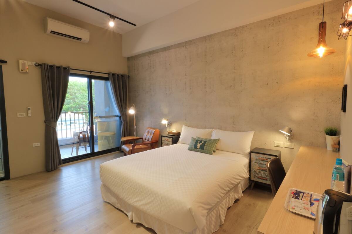 A modern hotel room with a large bed, a wooden desk, a chair, a small table, and a sliding glass door leading to an outdoor area.