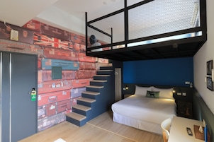 Una habitación de hotel compacta con una cama litera, un escritorio con silla, una mesita pequeña y un cuadro con motivos de equipaje.