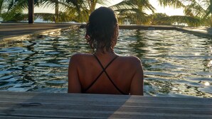 Una persona sentada junto a una piscina, con palmeras al fondo.