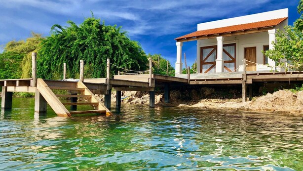 Un muelle de madera que conduce a un edificio blanco con techo rojo, ubicado junto a un cuerpo de agua con reflejos cristalinos.