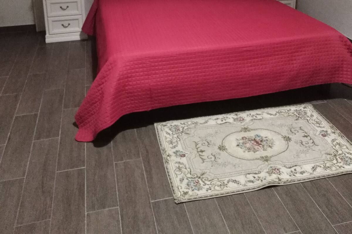 Un dormitorio con una cama, dos mesitas de noche, una mesita auxiliar, un cuadro colgado en la pared y una alfombra estampada.