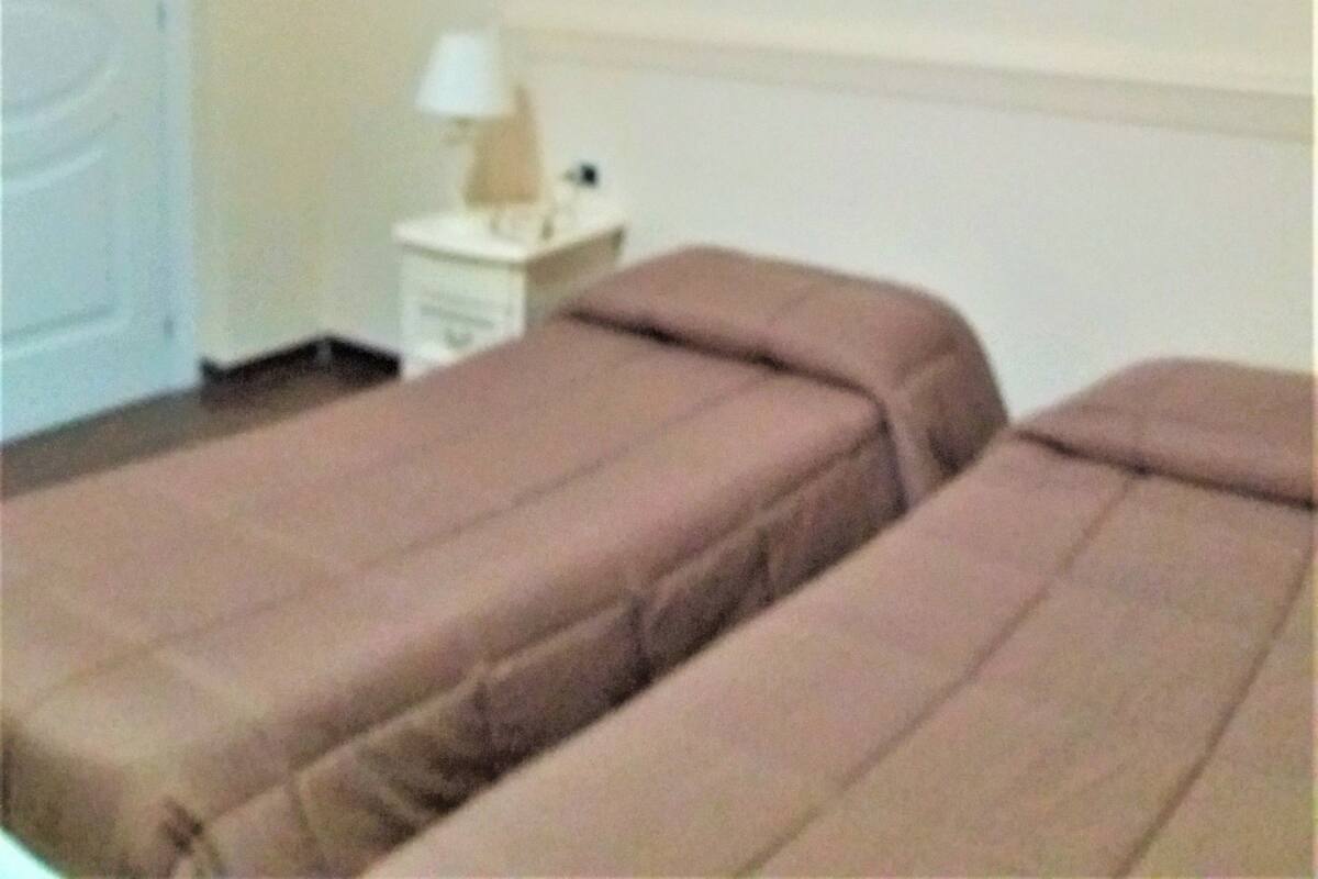 Una habitación con dos camas, una mesita de noche, una lámpara y un cuadro que representa una escena nocturna con columnas.