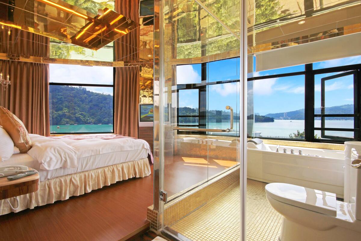 A bedroom with a bed, a glass-enclosed bathroom with a bathtub, and a view of a lake and mountains.