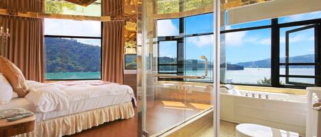 A bedroom with a bed, a glass-enclosed bathroom with a bathtub, and a view of a lake and mountains.