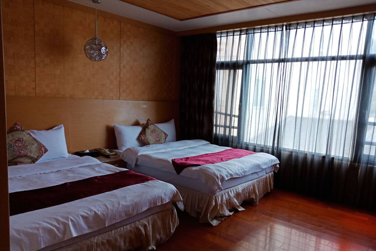 A hotel room with two beds, wooden flooring, a large window with sheer curtains, and a wooden ceiling.