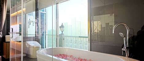 A modern bathroom with a freestanding bathtub filled with rose petals, a large window, and a visible cityscape view.